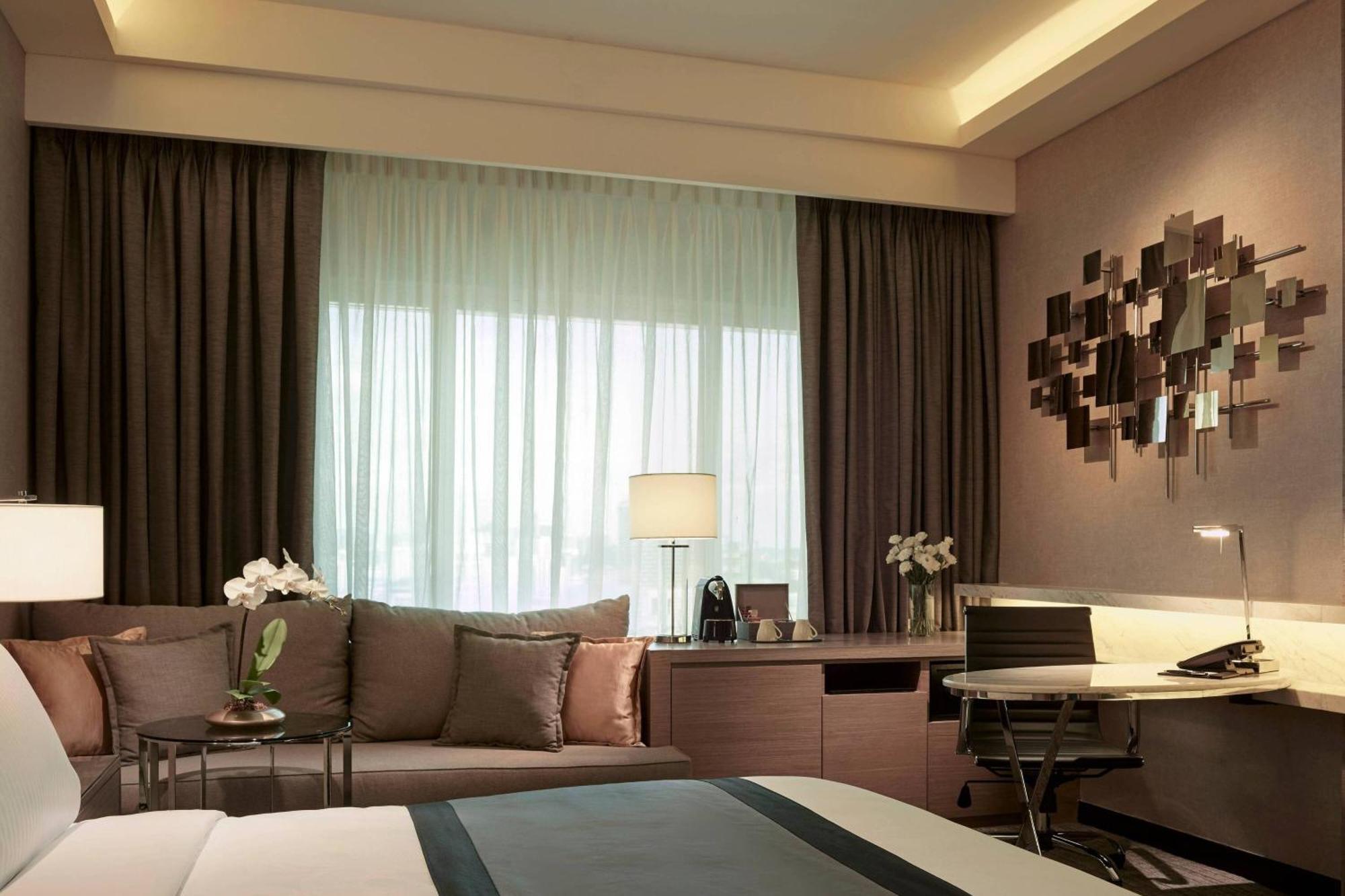 فندق جيه.دابليو ماريوت، كوالالمبور المظهر الخارجي الصورة The photo shows a modern hotel room interior. There's a comfortable-looking bed with a dark-colored bedspread. A sitting area features a sofa adorned with cushions next to a window, which is partially covered by light, sheer curtains. A lamp is place