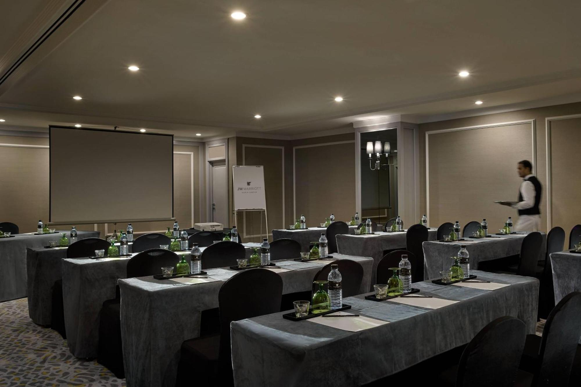 فندق جيه.دابليو ماريوت، كوالالمبور المظهر الخارجي الصورة The image shows a conference room setup. There are several tables arranged in a U-shape, each covered with a gray tablecloth. On each table, there are bottles of water and other supplies placed strategically. The room is softly lit with ceiling light